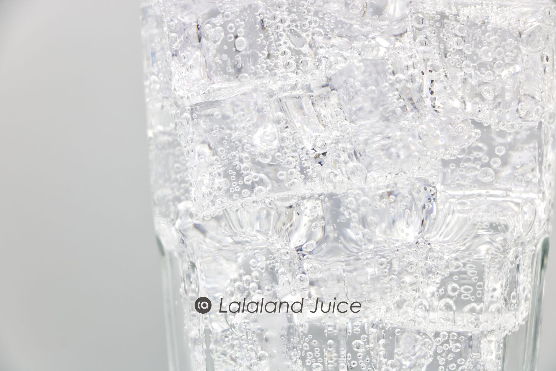 A tall glass packed with ice and sparkling water in close-up, beaded and cold — the Lalaland Juice mark set quietly within.