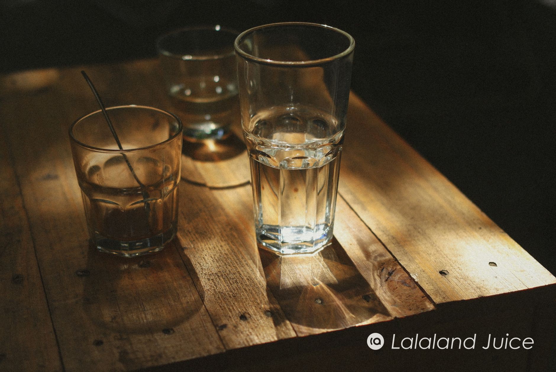 A tall glass of water catching a shaft of warm morning light on a wooden table — the first small kindness of a gentle morning — with the Lalaland Juice mark set within.